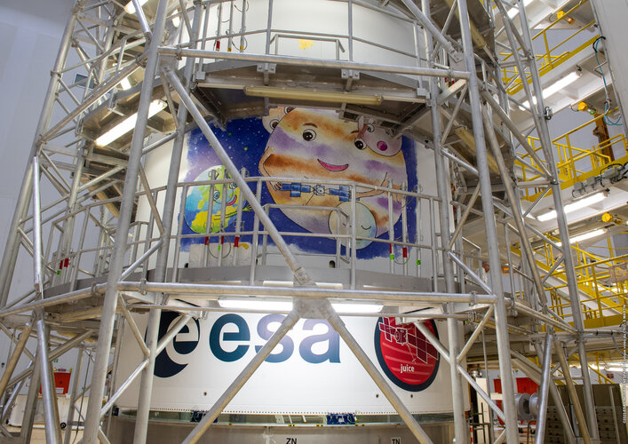 A close up of an Ariane 5 rocket surrounded by scaffolding. In the centre of the Ariane 5 is the sticker showing the artwork (blue background with Jupiter, three icy moons, Earth and Juice. All are smiling and Jupiter is holding Juice in its hands). Below the artwork is an ESA logo and the Juice mission patch (a round design with an outline of the spacecraft).