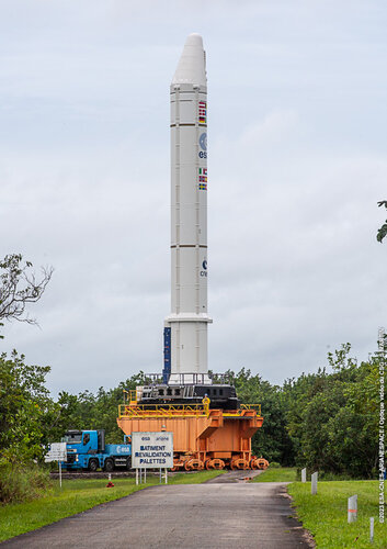 Launcher booster on top of a platform running along rails, pulled by a lorry. The image is taken outdoors, the sky is cloudy, in the foreground is a path and some trees